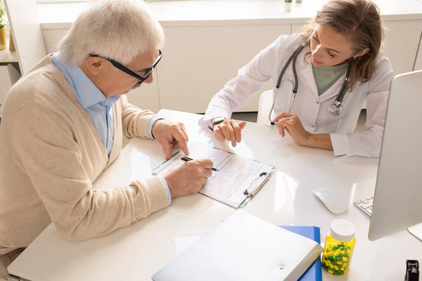 Senior Patient mit Stift zeigt auf ärztliches Dokument in Klemmbrett, während er seinem Arzt Fragen zu einem seiner Punkte in Kliniken stellt - Foto, Bild