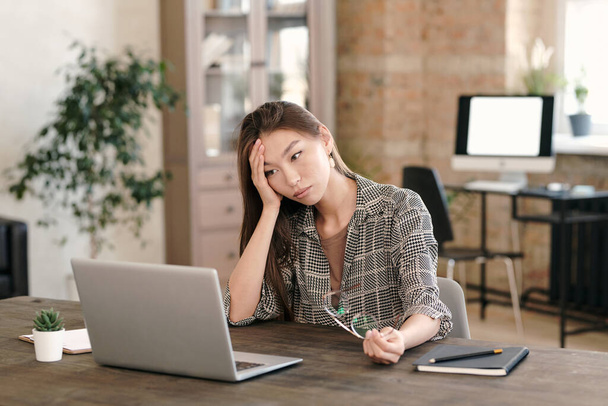 Ofis ortamında dizüstü bilgisayarın başında oturan aşırı çalışan ya da yorgun iş kadını kafasına dokunuyor. - Fotoğraf, Görsel