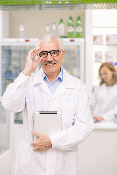 Succesvolle senior drogisterij werknemer in whitecoat en bril kijken naar u met een glimlach, terwijl het gebruik van digitale tablet tegen displays en collega - Foto, afbeelding