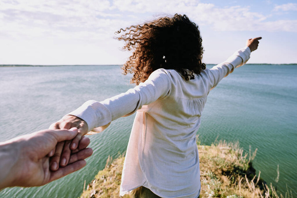Rear view of curly-haired girl holding hand of boyfriend and showing wonderful horizon at waterside, follow me concept - Zdjęcie, obraz