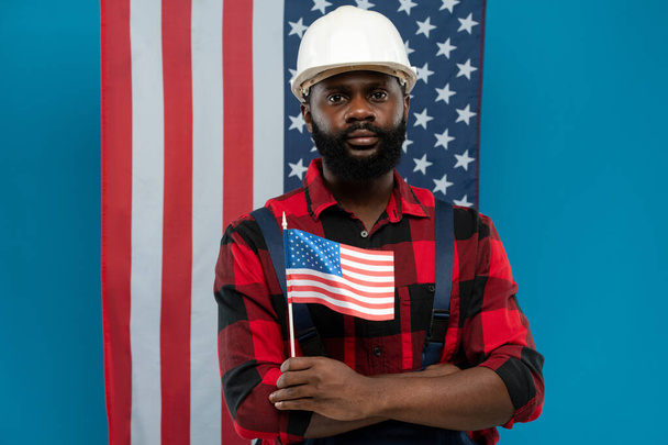 Junger bärtiger Mechaniker oder Reparateur in Flanell und Overalls mit kleiner amerikanischer Flagge vor Sternen-und-Streifen-Hintergrund - Foto, Bild