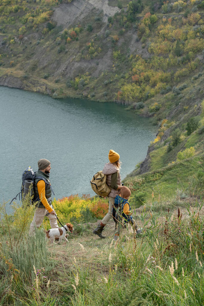 Současné Backpackers v teplém casualwear pohybující se po pěšině podél řeky nebo jezera a zároveň se těší víkendový výlet v přírodním prostředí - Fotografie, Obrázek