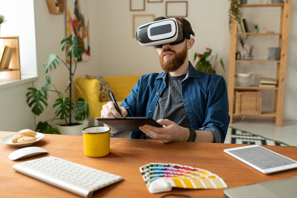 Young contemporary creative designer in vr headset looking at virtual display while drawing on tablet with stylus by workplace at home - Photo, Image