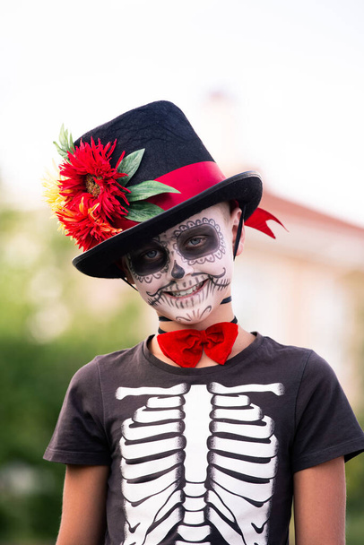 Netter Schuljunge mit bemaltem Gesicht im Halloween-Kostüm aus Skelett mit elegantem schwarzen Hut, der vor der Kamera steht und dich anlächelt - Foto, Bild