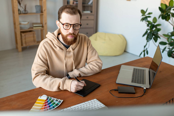 Jovem designer freelance masculino sério contemporâneo em óculos e casualwear usando touchpad e stylus enquanto retocando imagens - Foto, Imagem