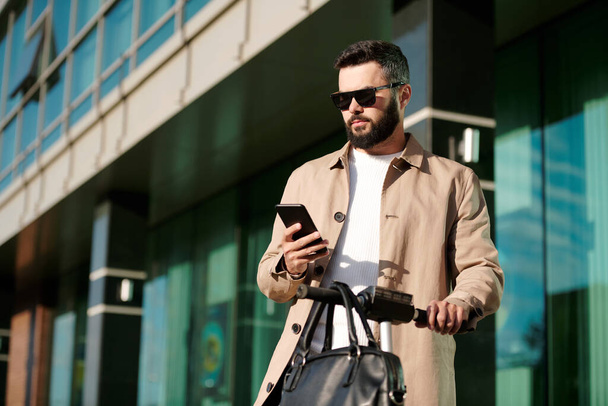 Σοβαρός νεαρός επιχειρηματίας σε μπεζ παλτό και γυαλιά ηλίου που κατέχουν από λαβές ηλεκτρικών σκούτερ, ενώ κύλιση σε εξωτερικούς χώρους smartphone - Φωτογραφία, εικόνα