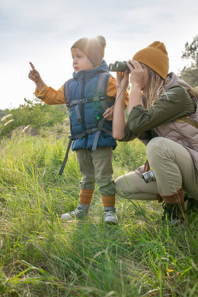 Молода жінка з біноклями дивиться, де її милий маленький син вказує під час поїздки на вихідні в природному середовищі серед зеленої трави
 - Фото, зображення