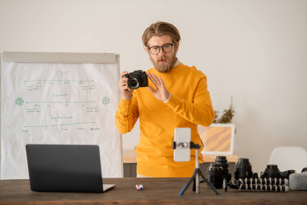 Young bearded man with photocamera explaining how to take stock photos to his online audience in front of smartphone camera - Photo, image