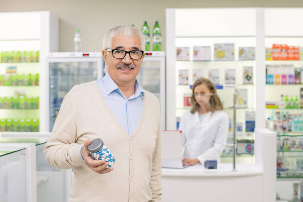 Happy aged man in casualwear en bril met grote fles pillen terwijl staande voor de camera tegen drogisterij assistent - Foto, afbeelding