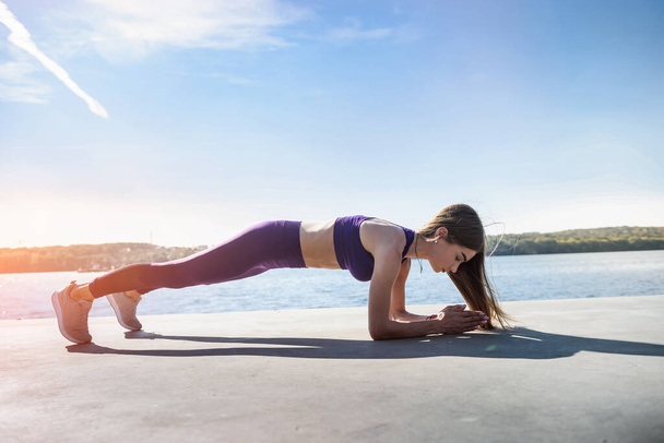 young female athlete doing push-ups outdoors near the lake at daytime. Healthy lifestyle - Photo, Image