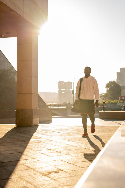 Jovem esportista ativo com capuz branco, tênis vermelho e calças pretas correndo para baixo pavimento de azulejos com longa sombra à sua esquerda - Foto, Imagem