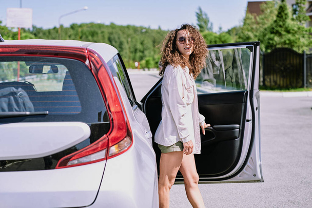Attractive curly-haired girl in sunglasses standing at open car door and looking at camera while traveling by car - Photo, Image