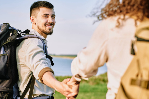 Sonriente joven mochilero barbudo cogido de la mano de su novia y mirándola mientras caminan juntos - Foto, Imagen