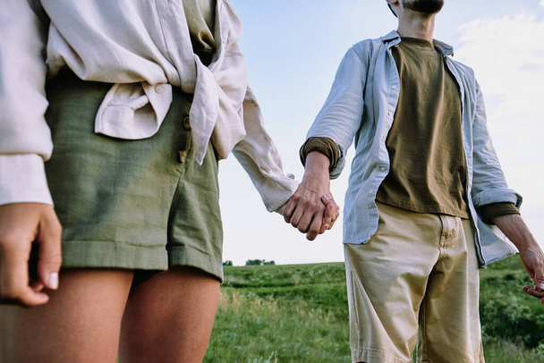 Nahaufnahme eines nicht wiederzuerkennenden reisenden Paares in lässigen Hemden, das Händchen haltend auf einem Feld steht - Foto, Bild