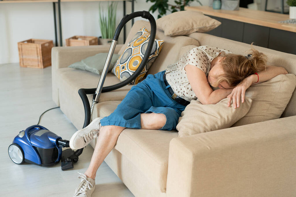 Mature exhausted blond female lying on soft pillows on couch while having rest after cleaning all the rooms in flat or house with vacuum cleaner - 写真・画像