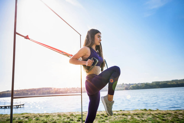 Joven mujer delgada haciendo entrenamiento de suspensión con correas de fitness al aire libre cerca del lago durante el día. Estilo de vida saludable - Foto, imagen