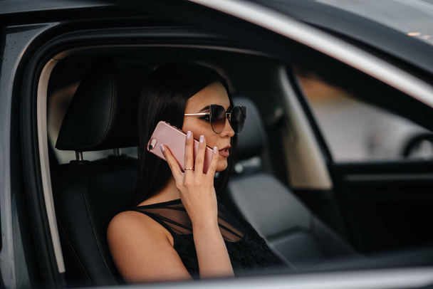 Stylish young girl sitting in a business class car in a black dress and talking on the phone. Business fashion and style - Fotó, kép