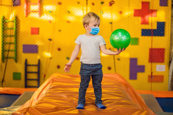 Little boy wearing a medical mask during COVID-19 coronavirus with a ball on trampolines - Photo, image