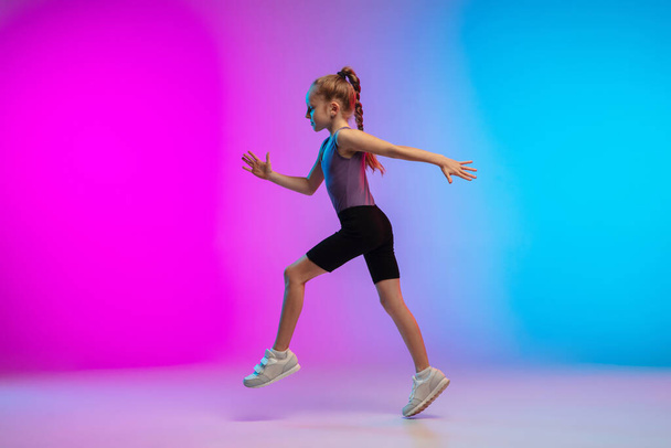 Teenage girl running, jogging against gradient pink-blue neon studio background in motion - Zdjęcie, obraz