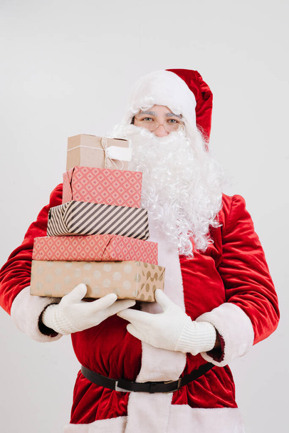 Joyous satisfied Santa Claus standing with hands full of wrapped gift boxes for Christmas. Over grey background. - Foto, Imagem