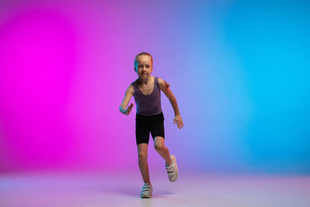 Teenage girl running, jogging against gradient pink-blue neon studio background in motion - Fotoğraf, Görsel
