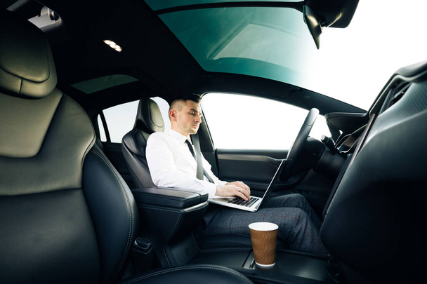 Self-driving electric car autopilot demanding driver attention to hold steering wheel and take control on highway. Businessman working on laptop at sunset. - Fotografie, Obrázek