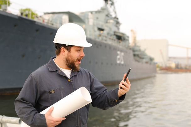 Морской инженер в шлеме держит рацию УКВ и бумаги рядом с судном на заднем плане. - Фото, изображение