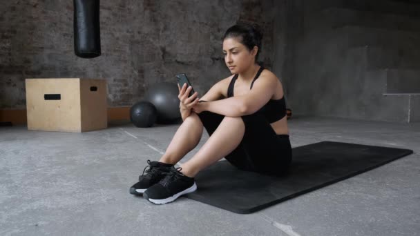 Tired woman is sitting on the mat in indoor gym, concentrating, deeply breathing and getting ready for. Girl, extreme. - Footage, Video