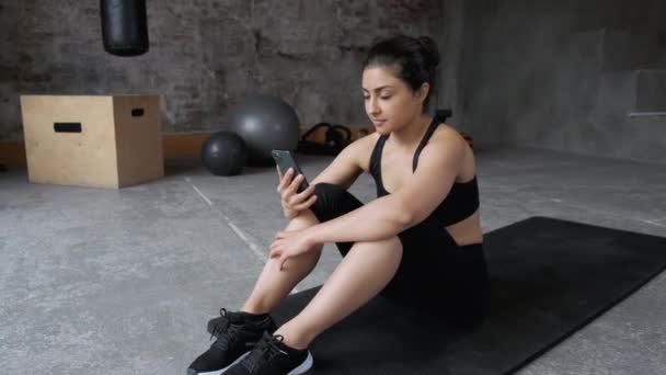 Una joven india usa un Smartphone para entrenar, vestida con ropa deportiva Leggings negros y un top está sentada cansada y descansando en el gimnasio - Metraje, vídeo