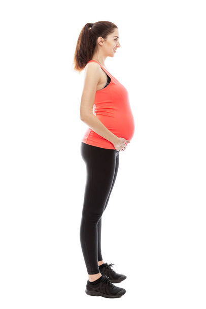A pregnant woman in sportswear is standing in full growth fashioning. Profile view. Isolated on white background. Vertical. - Foto, Imagen