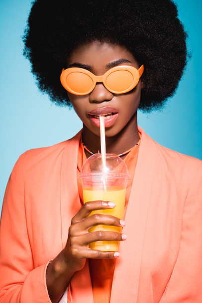 african american young woman in stylish outfit with orange juice isolated on blue background - Fotografie, Obrázek