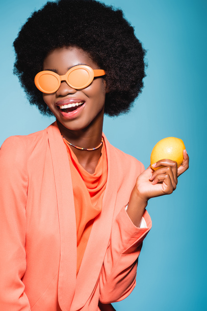 happy african american young woman in orange stylish outfit with lemon isolated on blue background - Foto, immagini