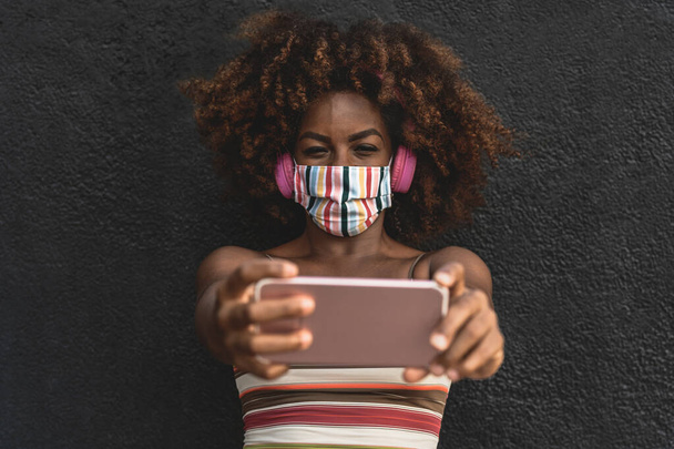 Mulher afro feliz tirar selfie com smartphone móvel enquanto vestindo máscara colorida rosto e ouvir música com fones de ouvido sem fio - Foto, Imagem