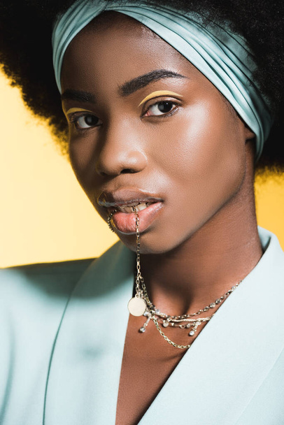 african american young woman in blue stylish outfit with necklace in mouth isolated on yellow - Foto, Bild