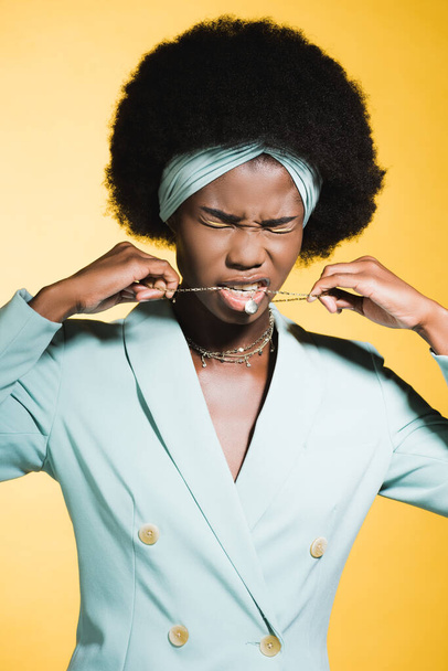 african american young woman in blue stylish outfit with necklace in mouth isolated on yellow - 写真・画像