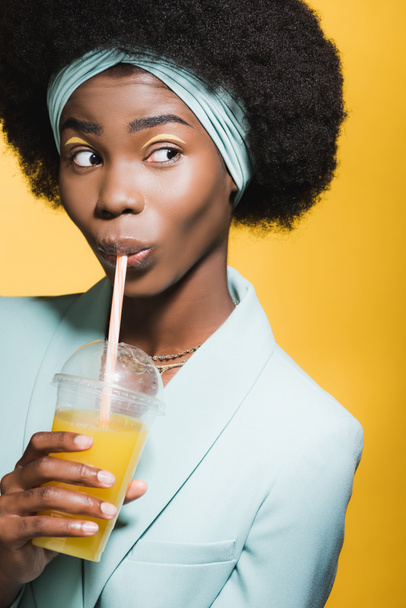 african american young woman in blue stylish outfit with orange juice isolated on yellow  - Фото, изображение