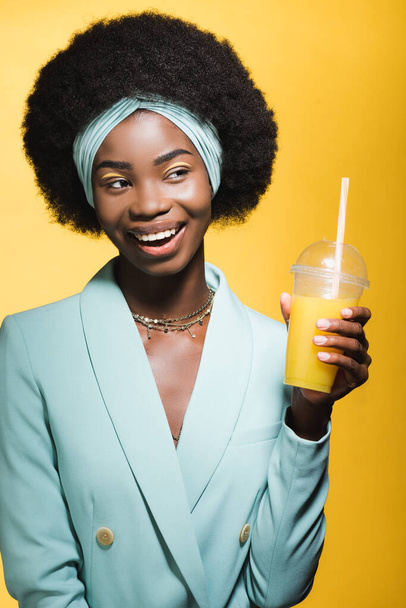 smiling african american young woman in blue stylish outfit with orange juice isolated on yellow  - Fotografie, Obrázek