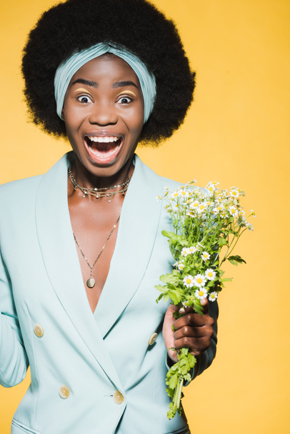 happy african american young woman in blue stylish outfit with chamomile bouquet isolated on yellow  - Фото, изображение
