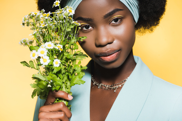 african american young woman in blue stylish outfit with chamomile bouquet isolated on yellow  - Foto, afbeelding