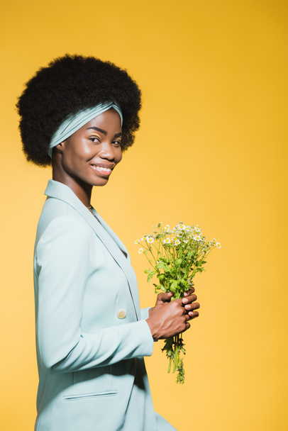 side view of smiling african american young woman in blue stylish outfit with chamomile bouquet isolated on yellow  - Foto, Imagem