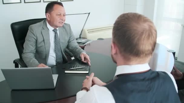 Plan grand angle de jeune homme avec barbe sur le visage assis au bureau devant son patron adulte mature l'écoutant - Séquence, vidéo