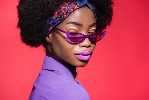 african american young woman in purple stylish outfit and sunglasses isolated on red - Фото, изображение