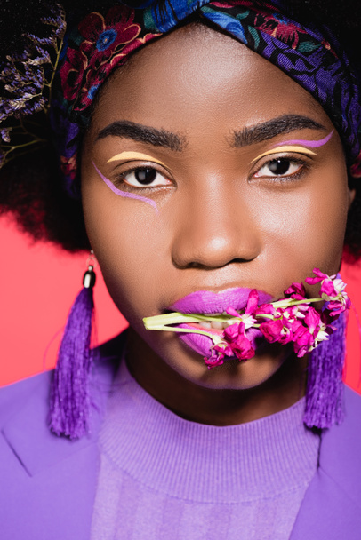 african american young woman in purple stylish outfit with flower in mouth isolated on red - Foto, imagen