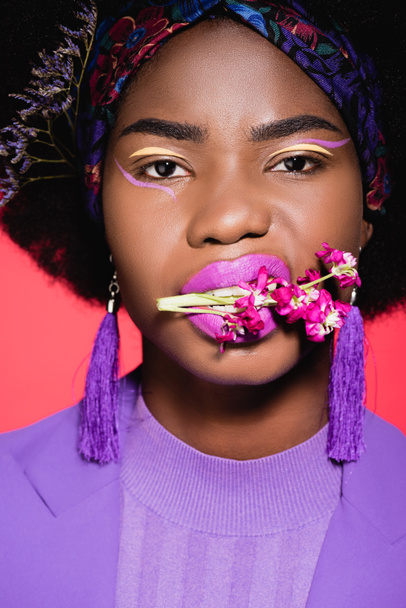 brutal african american young woman in purple stylish outfit with flower in mouth isolated on red - Foto, imagen