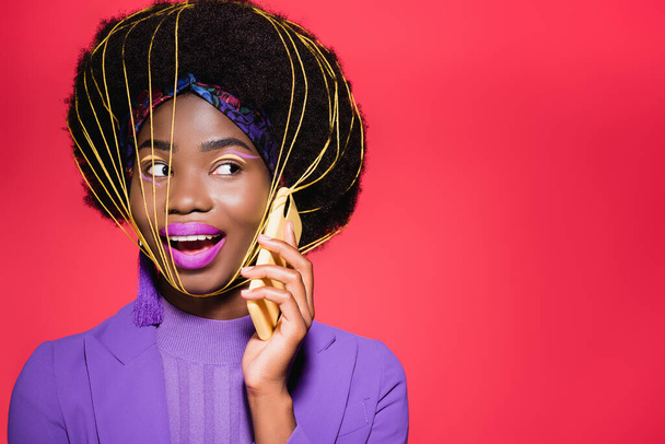 happy african american young woman in purple stylish outfit with yellow strings on face using smartphone isolated on red - Photo, Image