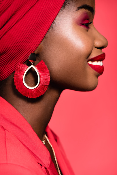 profile of smiling african american young woman in stylish outfit and turban isolated on red - Foto, Imagen