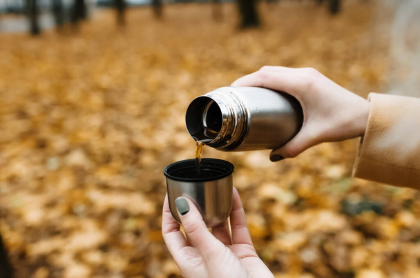 Woman pours tea from a thermos, outdoors. Close-up of female hands and a mug with tea. Autumn background. - Fotó, kép