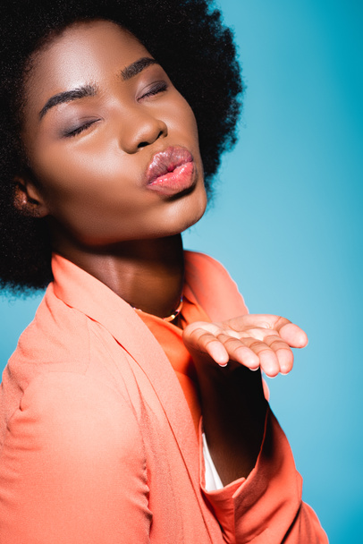 Afrikaans amerikaanse jonge vrouw in oranje stijlvolle outfit blazen kus geïsoleerd op blauw - Foto, afbeelding
