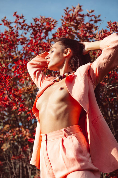 Young beautiful brunette girl with good makeup on her face with expressive features and figure. walks in the fall among beautiful red trees, she is wearing a stylish pink suit and she is a little naked - Foto, Bild