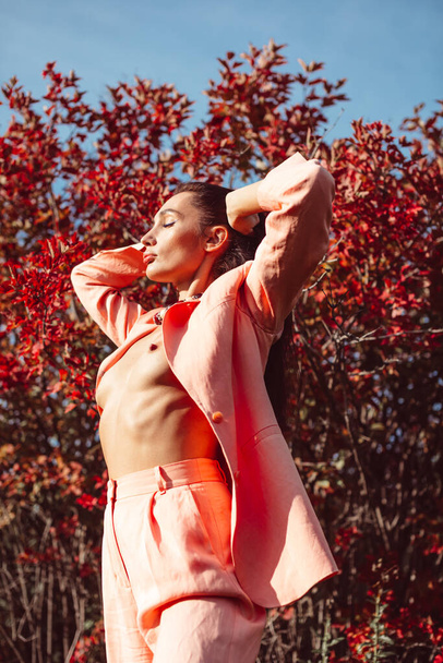 Young beautiful brunette girl with good makeup on her face with expressive features and figure. walks in the fall among beautiful red trees, she is wearing a stylish pink suit and she is a little naked - Foto, Imagen